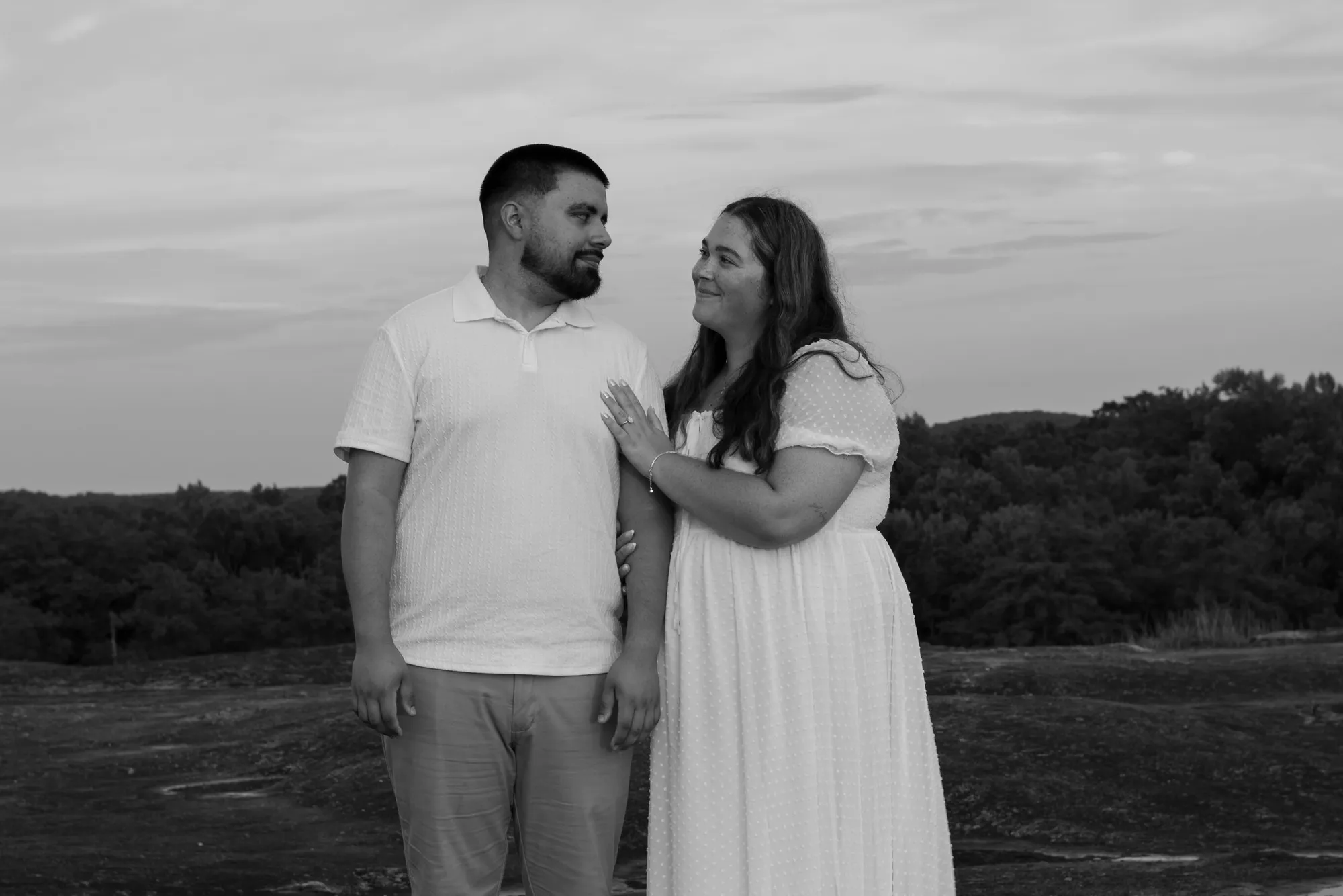 A couple on a granite overlook, she looks up at him as he smiles toward the camera, in black and white