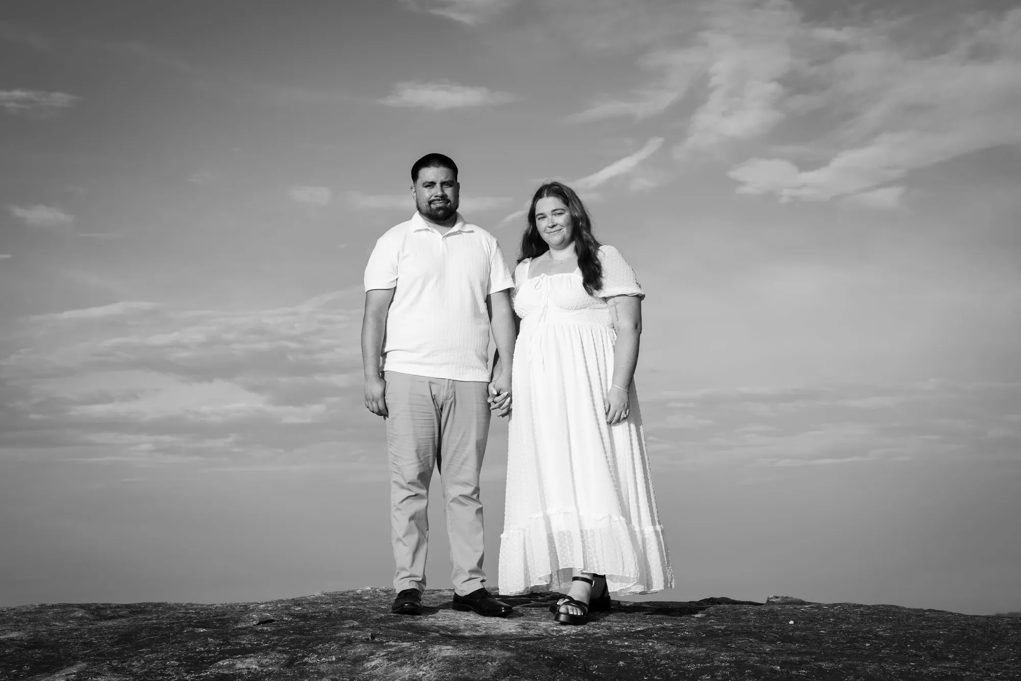 A couple standing hand in hand on a granite summit under a streaked sky, in black and white