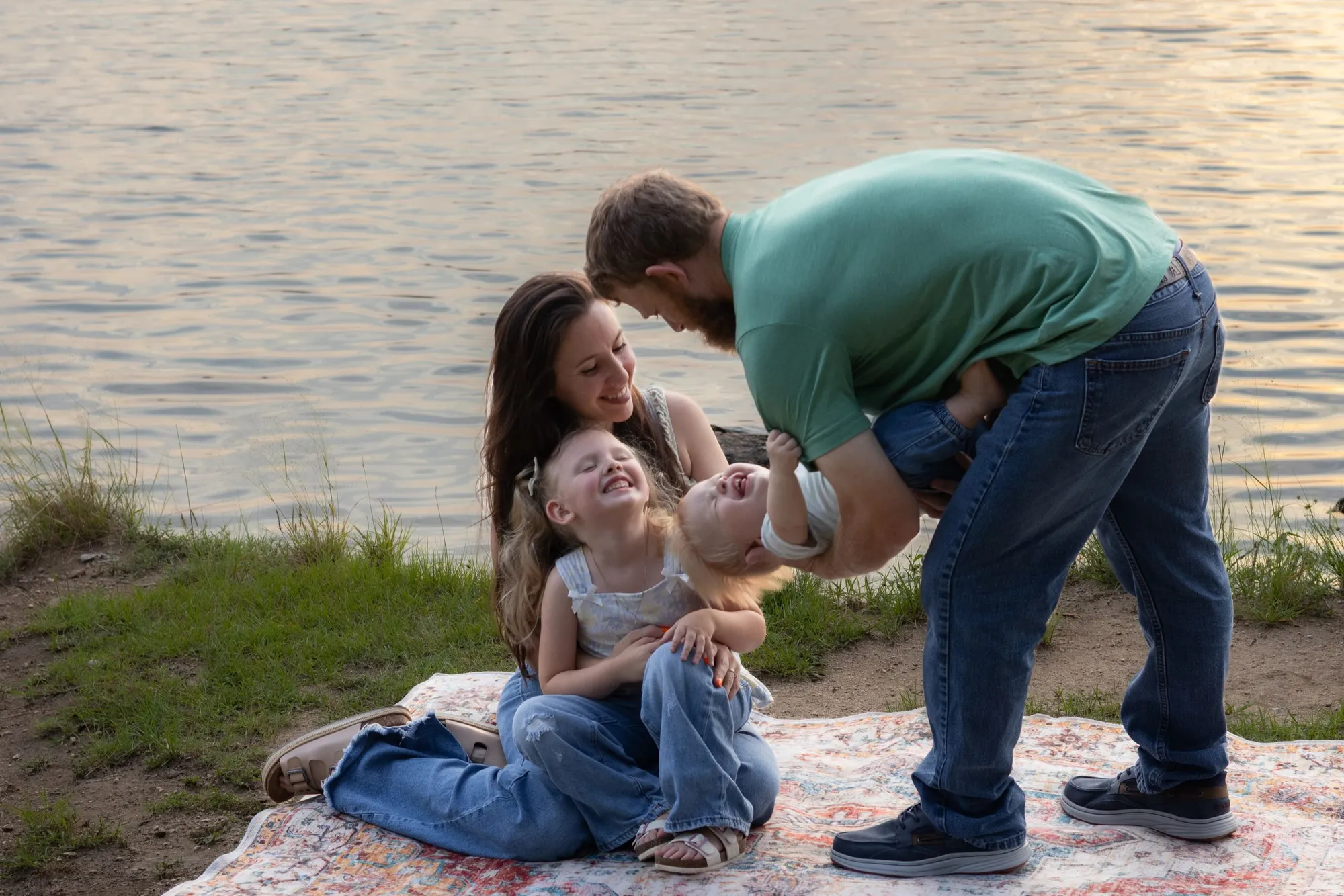 A mother and father playing with their two small children on a patterned blanket beside a lake