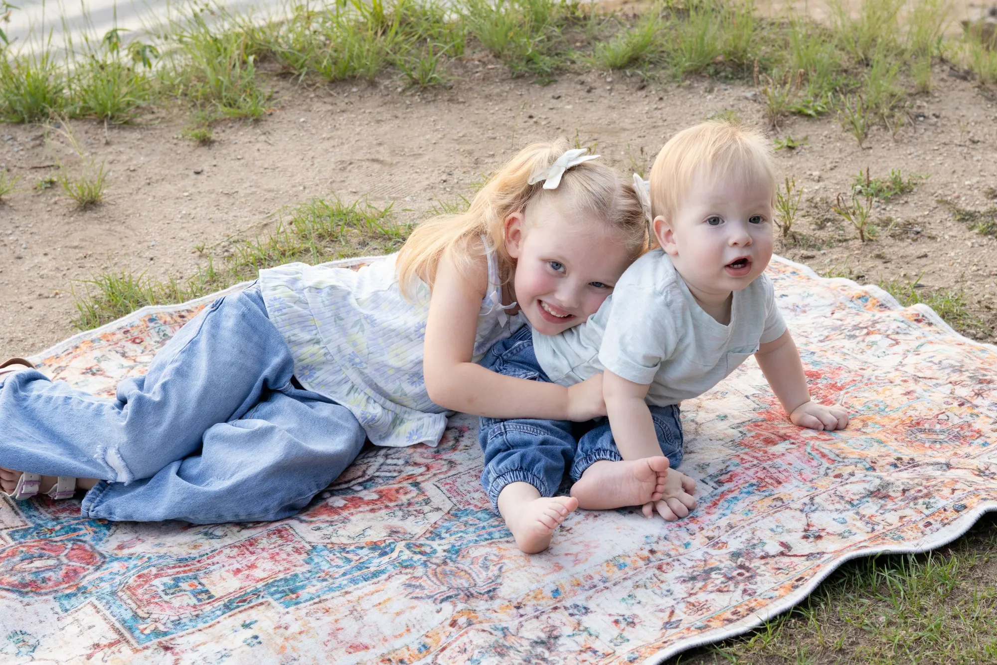 A little girl and her baby brother lying close together on a patterned rug in the grass