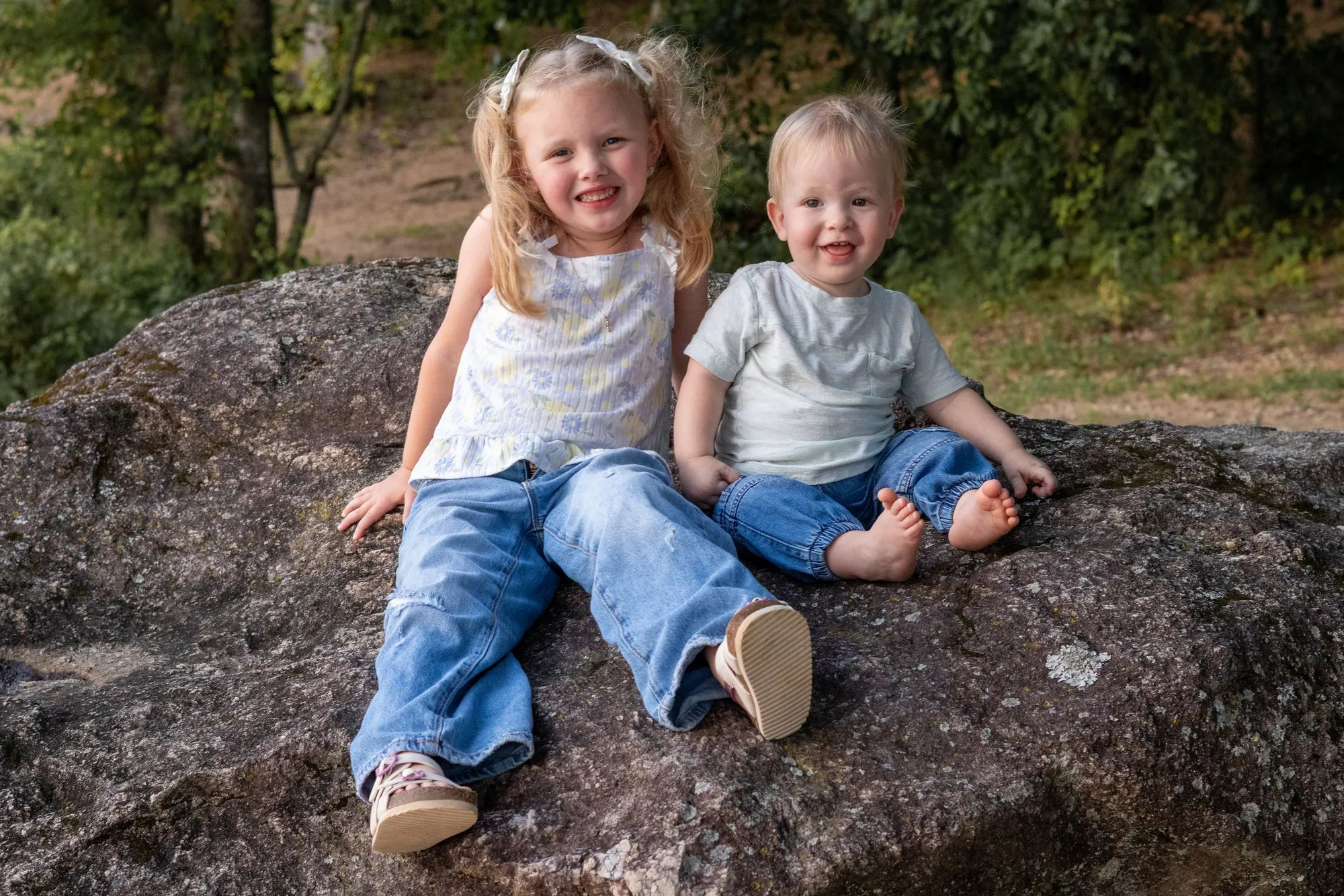 A little girl and her baby brother sitting side by side on a large boulder