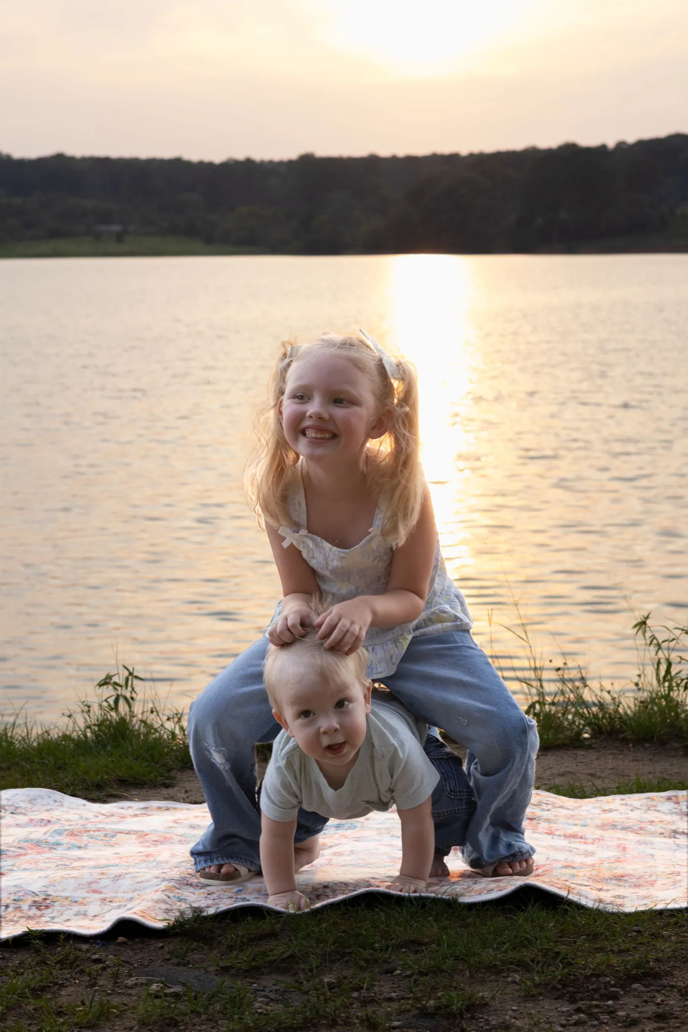 A little girl leaning over her crawling baby brother on a blanket, the sun setting over a lake behind them